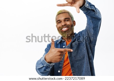Snap. Stylish young man takes picture of you, looks through finger frames and smiles happy, searching for perfect angle, picturing smth, stands over white background