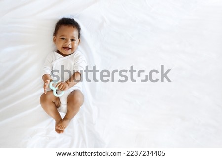 Childcare Concept. Portrait Of Adorable Little Black Baby Lying On Bed With Teether In Hand, Top View Shot Of Cute African American Infant Boy Or Girl Wearing Bodysuit Smiling At Camera, Copy Space Royalty-Free Stock Photo #2237234405