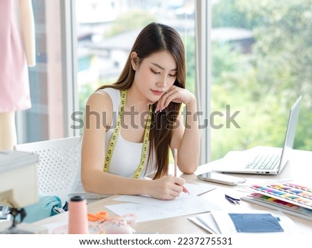 Millennial Asian young beautiful professional female dressmaker designer seamstress using pencil drawing sketching designing trendy style outfit on working desk in tailor workshop studio office.