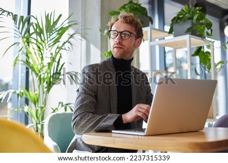 Portrait of young attractive businessman wearing stylish eyeglasses and suit using laptop computer working online sitting at workplace. Successful business 