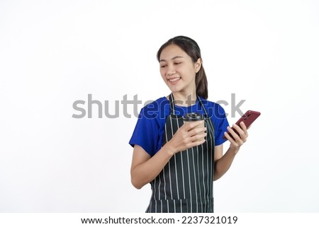 Excited asian coffee shop SME owner asian woman wearing blue t-shirt and black apron isolated on white background. Using phone and holding coffee cup