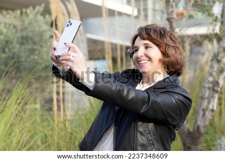 Smiling woman takes selfie on smartphone. Brunette, caucasian. Outside. City. Sunny autumn day. Concept of activity in social networks, mobile photo, lifestyle. Happy woman 40+. Selective focus.