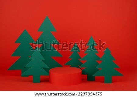 Festive Christmas background with red cylinder podium mockup, green paper spruces in art cartoon style for presentation cosmetic product, gifts, goods, copy space. New Year template for advertising.