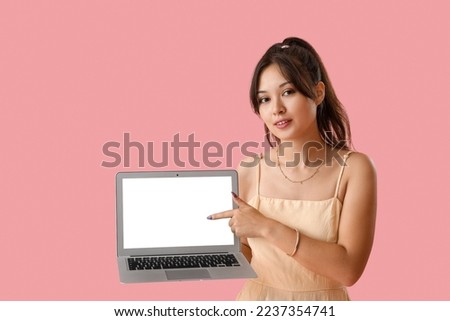 Young Asian woman pointing at laptop on pink background