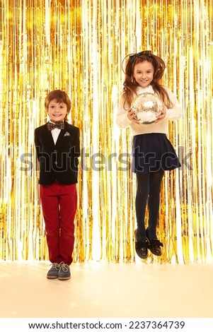 Kid's fashion and holidays. Joyful boy and girl in classical evening clothes pose merrily standing back-to-back on shiny gold foil curtains background. Children's party.