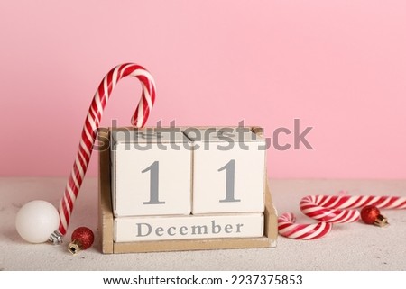 Cube calendar with date 11 DECEMBER, candy canes and Christmas balls on table near pink wall