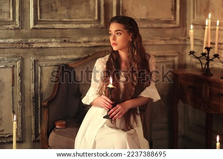 Young long haired woman in vintage night gown sitting on chair with candle in hands. Historic portrait of noble woman Royalty-Free Stock Photo #2237388695