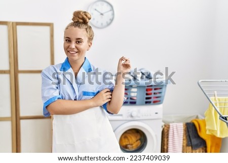 Young caucasian woman wearing cleaner uniform at the laundry room with a big smile on face, pointing with hand and finger to the side looking at the camera. 