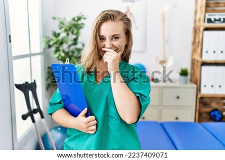 Young caucasian woman working at pain recovery clinic laughing and embarrassed giggle covering mouth with hands, gossip and scandal concept  Royalty-Free Stock Photo #2237409071