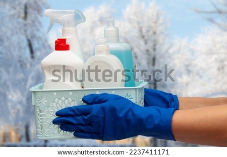 Woman's hands in rubber protective gloves holding plastic basket with bottles of glass and tile cleaner with winter landscape on the background. Washing and cleaning concept.