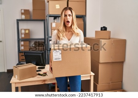 Young blonde woman working at small business ecommerce holding big box in shock face, looking skeptical and sarcastic, surprised with open mouth 