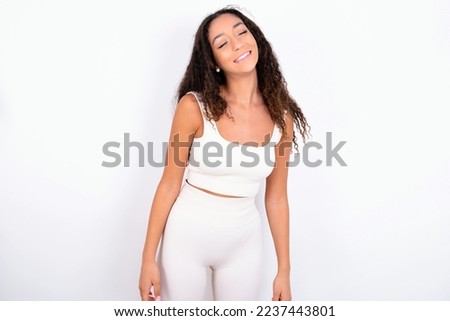 Positive teen girl with curly hair wearing white sport set over white background with overjoyed expression closes eyes and laughs shows white perfect teeth Royalty-Free Stock Photo #2237443801