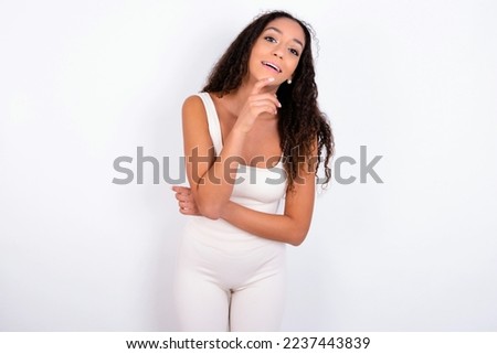teen girl with curly hair wearing white sport set over white background laughs happily keeps hand on chin expresses positive emotions smiles broadly has carefree expression Royalty-Free Stock Photo #2237443839