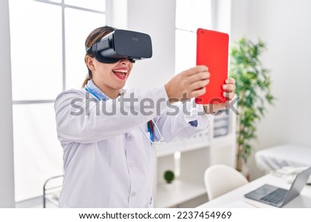 Young beautiful hispanic woman doctor using virtual reality glasses and touchpad at clinic