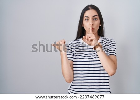 Young brunette woman wearing striped t shirt asking to be quiet with finger on lips pointing with hand to the side. silence and secret concept. 
