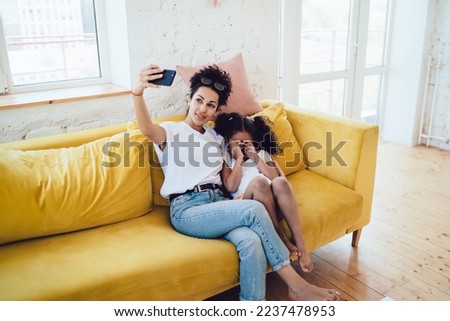 Cheerful smiling African American young woman taking selfie with curly adorable daughter hiding face from camera with hands on yellow cozy couch at home