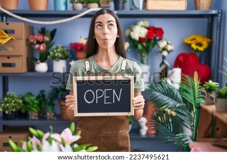 Young brunette woman working at florist holding open sign making fish face with mouth and squinting eyes, and comical. 