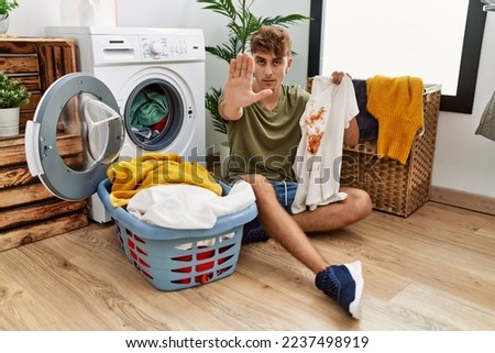 Young caucasian man doing laundry holding t shirt with stain with open hand doing stop sign with serious and confident expression, defense gesture 