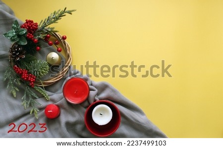 On a yellow background, a gray plaid with a wreath with fir branches and holly berries, candles and the text "2023" denoting the coming year. Flat Lay on the eve of the New Year and Merry Christmas