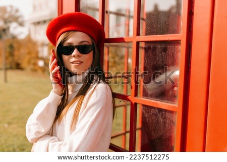 Close up outside photo of stylish european girl with dark loose hair wearing sunglasses, red beret and white shirt talking on smartphone. 