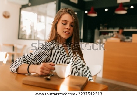 Close up indoor portrait of young attractive pretty girl with long hair wearing striped shirt sitting in sunny cafe in morning and enjoying coffee and croissant.