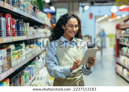 Latin American woman seller happy and smiling uses tablet computer in supermarket to check stock availability, shop assistant among shelves with goods in apron. Royalty-Free Stock Photo #2237513183