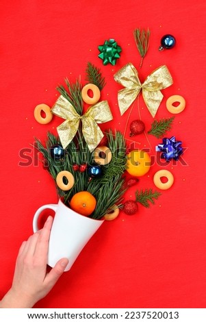 Christmas decor pouring out of a white cup on a red background. Cover postcard for the New Year, the mood of the holiday and fun. Woman's hand holding a cup with Christmas decorations