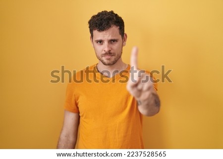 Young hispanic man standing over yellow background pointing with finger up and angry expression, showing no gesture 