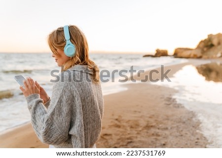 Beautiful young woman wearing sweater listening music with headphones and mobile phone while standing at sunny beach Royalty-Free Stock Photo #2237531769