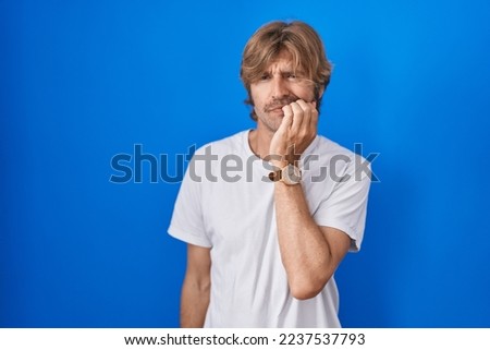 Middle age man standing over blue background looking stressed and nervous with hands on mouth biting nails. anxiety problem. 