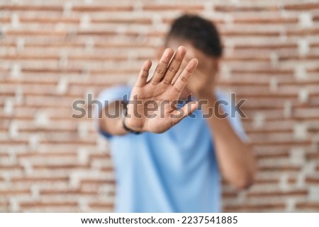 Brazilian young man standing over brick wall covering eyes with hands and doing stop gesture with sad and fear expression. embarrassed and negative concept. 