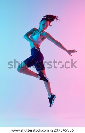Portrait of young sportive woman training, doing exercises, jumping isolated over gradient blue pink background in neon light. Concept of sport, strength, body care, fitness, wellbeing, health. Ad
