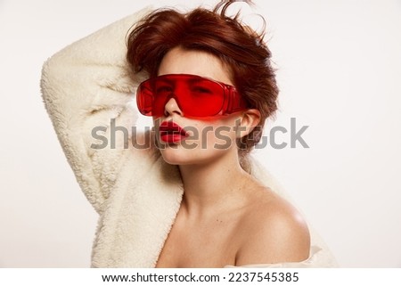Portrait of young beautiful red-haired woman posing in fur coat and red trendy glasses isolated over white background. Cosmetological treatment. Concept of hight fashion, make-up, magazine style, ad