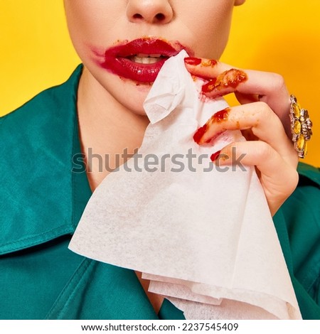 Cropped image of woman in green coat with smudged red lipstick cleaning mouth after eating nuggets over yellow background. Food pop art photography. Complementary colors. Copy space for ad, text