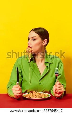 Young girl eating spaghetti, noodles sticking out of the mouth over yellow background. Dinner time. Italian food lover. Food pop art photography. Complementary colors. Copy space for ad, text