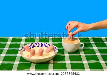 Plate with mashed potatoes and sausages on green tablecloth over blue background. Woman dipping sausage into coffee. Food pop art photography. Complementary colors. Copy space for ad, text