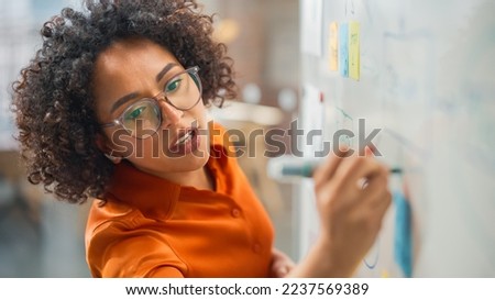 Modern Creative Agency Meeting: Confident Black Female Engineer Uses Whiteboard, Makes Report to a Group of Engineers, Managers Talks, and Shows Statistics, Growth and Analysis Information Royalty-Free Stock Photo #2237569389
