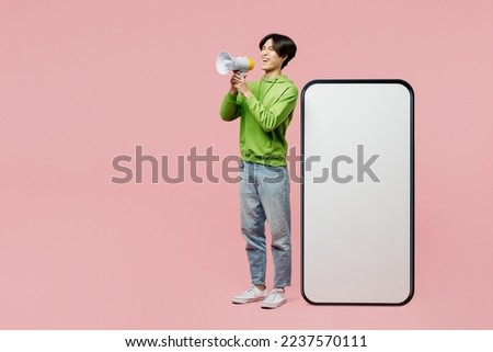 Full body excited young man of Asian ethnicity wear green hoody big huge blank screen mobile cell phone smartphone with area scream in megaphone isolated on plain pastel light pink background studio