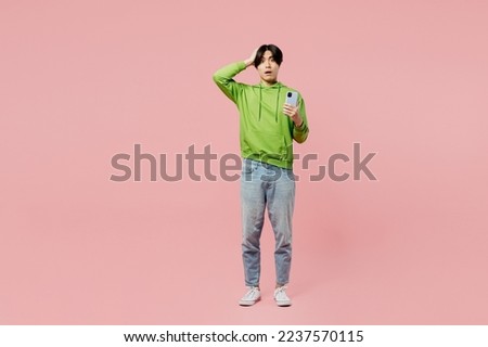 Full body shocked sad young man of Asian ethnicity wear green hoody look camera use mobile cell phone hold head isolated on plain pastel light pink background studio portrait. People lifestyle concept Royalty-Free Stock Photo #2237570115