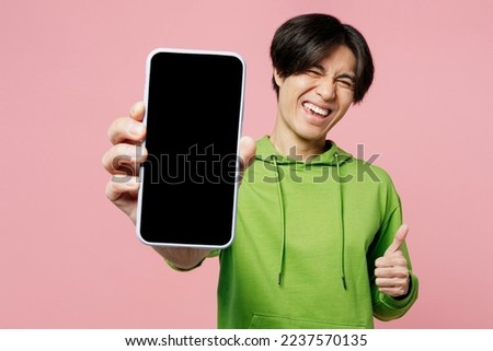 Young fun man of Asian ethnicity wear green hoody look camera hold in hand use close up mobile cell phone with blank screen workspace area show thumb up isolated on plain pastel light pink background Royalty-Free Stock Photo #2237570135