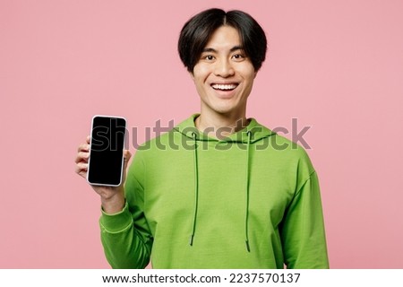 Young man of Asian ethnicity wear green hoody look camera hold in hand use mobile cell phone with blank screen workspace area isolated on plain pastel light pink background. People lifestyle concept
