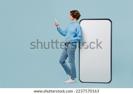 Full body happy side view young woman wear knitted sweater near big huge blank screen mobile cell phone with workspace mockup area use smartphone isolated on plain pastel light blue cyan background
