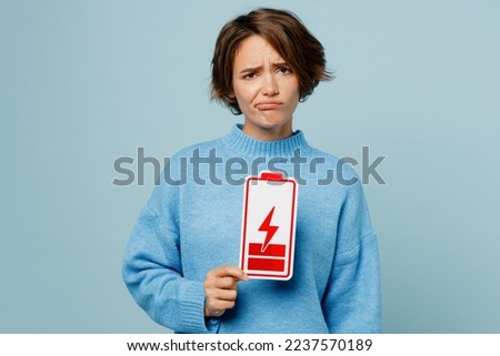 Young weary sad caucasian woman wear knitted sweater look camera hold in hand low battery red card sign isolated on plain pastel light blue cyan background studio portrait. People lifestyle concept Royalty-Free Stock Photo #2237570189