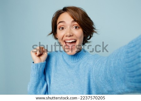 Close up young hapy caucasian woman wear knitted sweater look camera doing selfie shot pov on mobile cell phone do winner gesture isolated on plain pastel light blue cyan background studio portrait