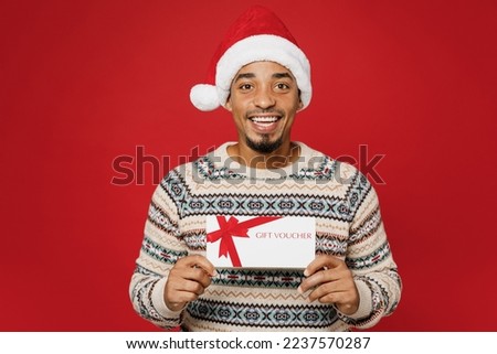 Merry young man wear warm cozy Christmas sweater Santa hat posing hold gift certificate coupon voucher card for store isolated on plain red background. Happy New Year 2023 celebration holiday concept