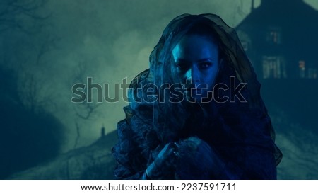 Beautiful witch making the witchcraft over the smoky background at night. Scary house on the hill. Halloween image.
