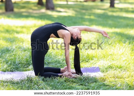 Young pregnant woman leading a healthy lifestyle and practicing yoga, performs a variation of the ushtrasana exercise, camel pose, trains in the park in a black sports overalls