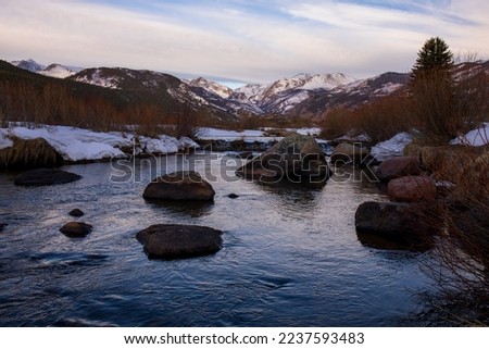Snow covered river banks in nature setting with a slow flowing stream and a pale sky.  Rocky Mountain stream with blue water, dormant grass.  Colorado high country with cloud filled sunrise sky. Royalty-Free Stock Photo #2237593483