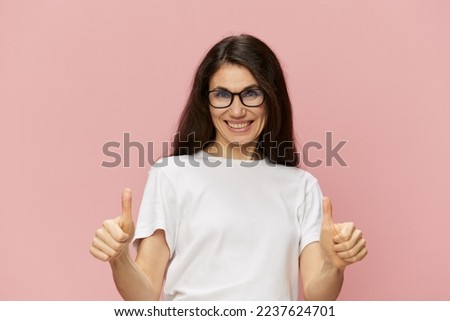 portrait of a beautiful, smart, pleasant woman in a light T-shirt and black-rimmed glasses smiling, raising her thumb up. Horizontal photo on a pink background with empty space
