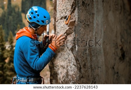 Man with climbing equipment climbs the cliff to the top against the backdrop of a beautiful valley with a forest. A tourist in a helmet and with a hammer, is engaged in mountaineering, extreme sports.